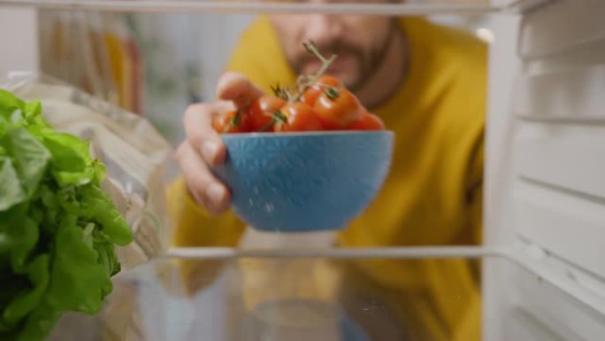 厨房冰箱内的摄像头: 英俊的男人打开冰箱门，看着里面拿出一碗西红柿。准备健康餐的人。从装满健康食品的