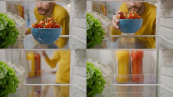 厨房冰箱内的摄像头: 英俊的男人打开冰箱门，看着里面拿出一碗西红柿。准备健康餐的人。从装满健康食品的