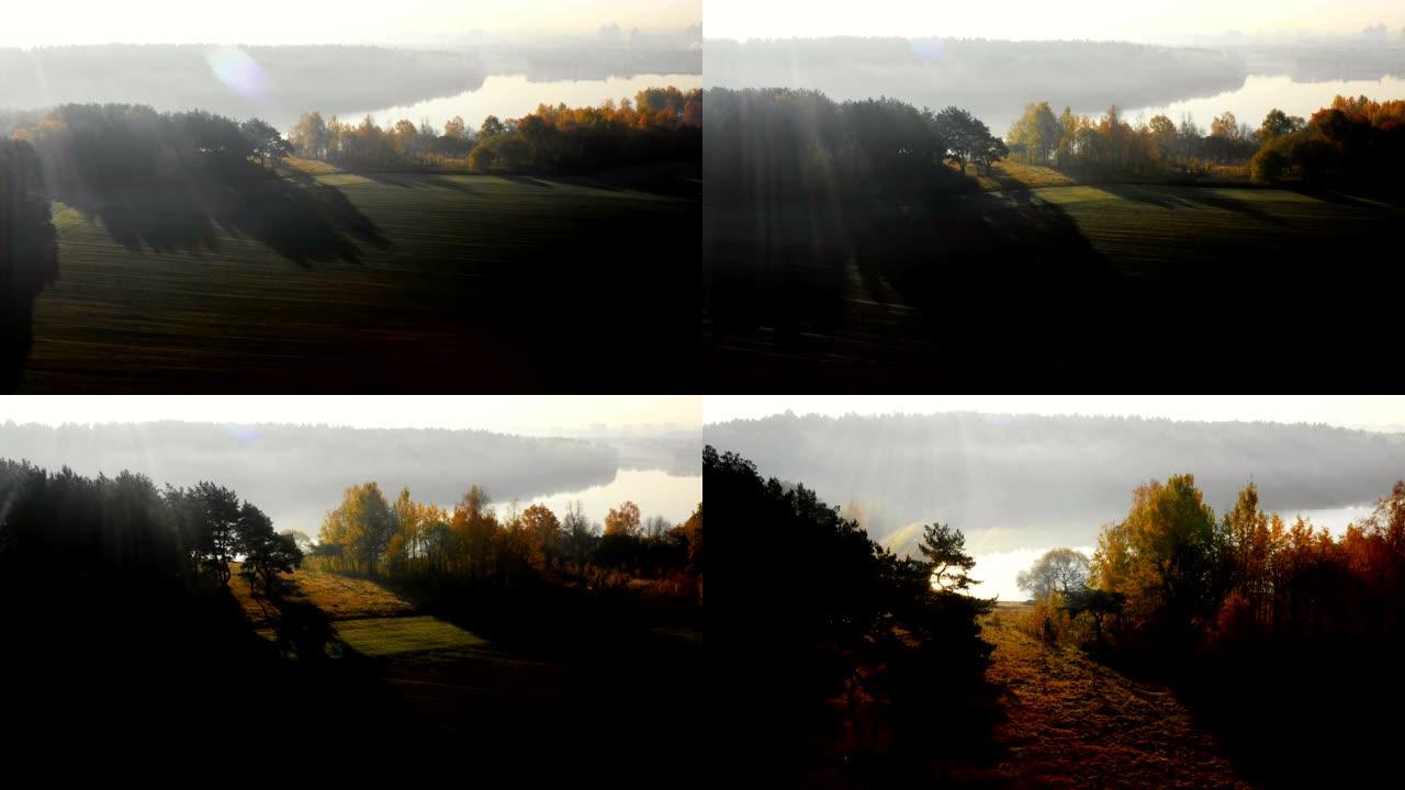 无人机降落在充满薄雾的秋日晴朗的田野和森林上，覆盖着日出雾，朝着小湖路。