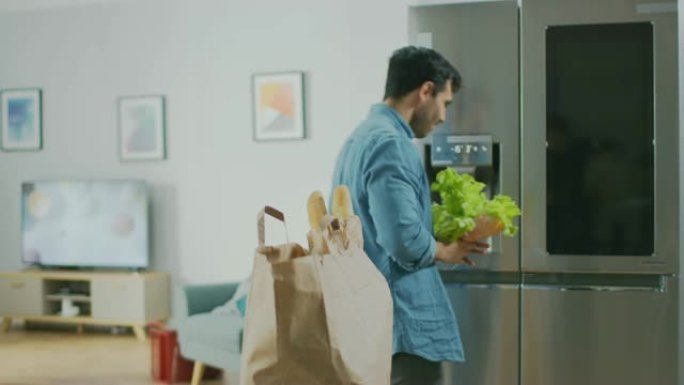 英俊迷人的年轻人把杂货带到厨房。他将新鲜的沙拉蔬菜放入现代冰箱中。他以轻盈的方式唱歌和跳舞。房间有明