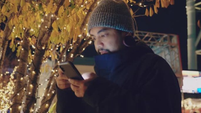 年轻的亚洲男子正站在智能手机上发短信，背景是圣诞灯。