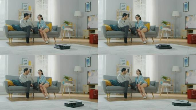 智能机器人真空吸尘器从地毯上吸尘的照片。美丽的夫妇坐在沙发上，在后台聊天。科技家电设备超越了它们。