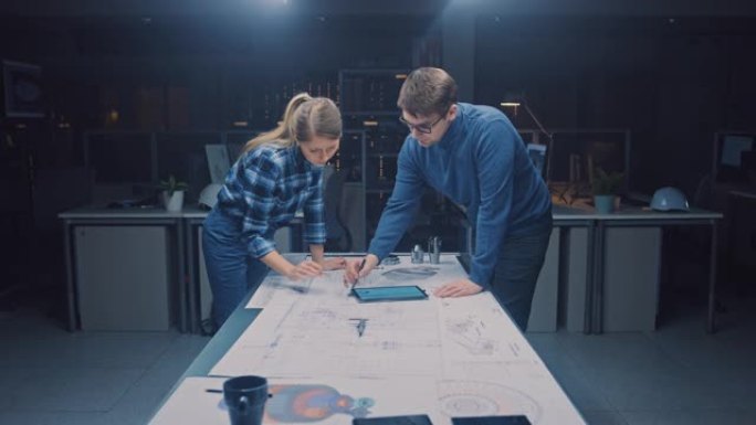 在黑暗的工业设计工程设施中，男女工程师使用会议桌交谈和工作蓝图。在桌面图纸、草稿和电动发动机部件、零