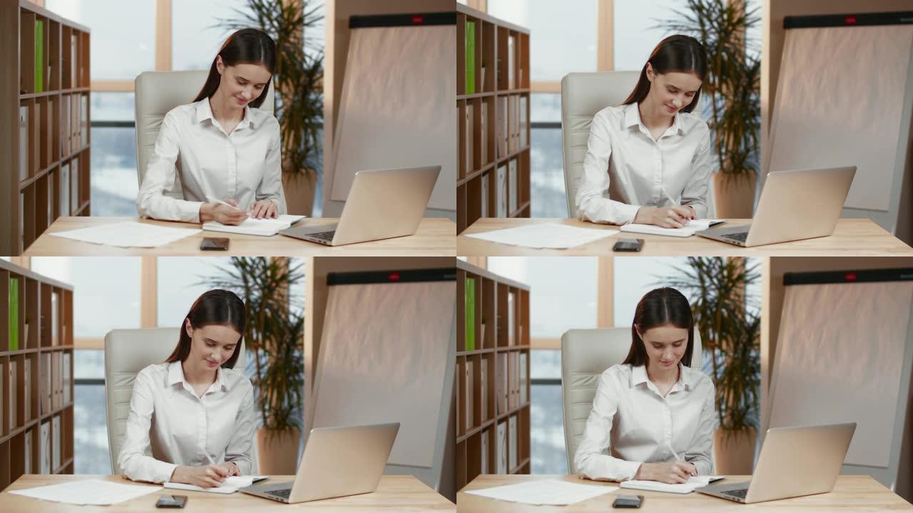 自信的女企业家计划她的工作日坐在办公桌前，拿着笔在办公室里看着笔记本