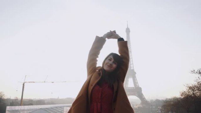 美丽快乐微笑的旅游女人的肖像坐着，在巴黎埃菲尔铁塔日出时举手慢动作。