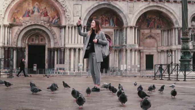 快乐兴奋的女性游客，鸽子坐在手臂上，在威尼斯慢动作的城市广场上拍摄智能手机照片。
