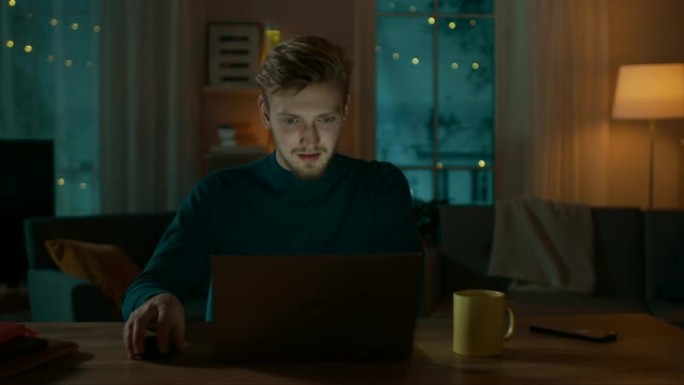 晚上在家: 英俊男子坐在办公桌前在笔记本电脑上工作的肖像。微笑的自由职业者在电脑上工作。