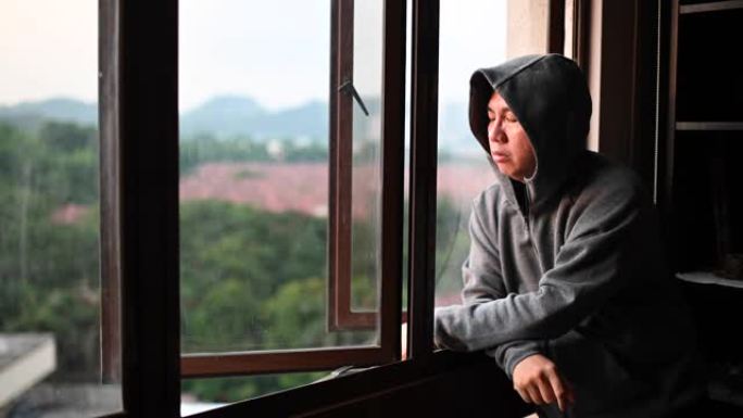 日落时分，一名身穿连帽衫的亚洲华人中年男子从家中望向窗外，表情严肃
