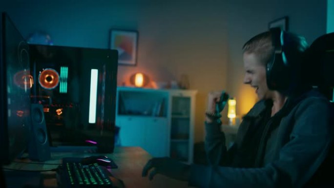 快乐的玩家在他的个人电脑上玩并赢得第一人称射击游戏在线视频游戏。房间和电脑有彩色霓虹灯。年轻人戴着耳