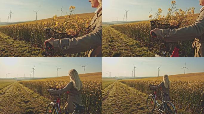 SLO MO女人在远处用风力涡轮机沿着油菜籽的田野推自行车