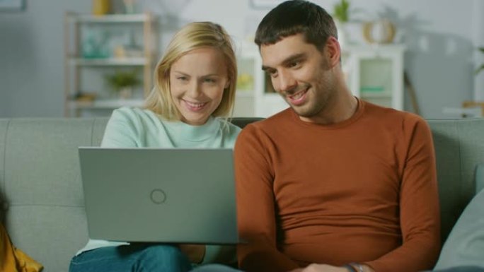 可爱的年轻夫妇坐在家里的沙发上，他们看起来用笔记本电脑，玩得很开心。美丽幸福的年轻夫妇恋爱的肖像。