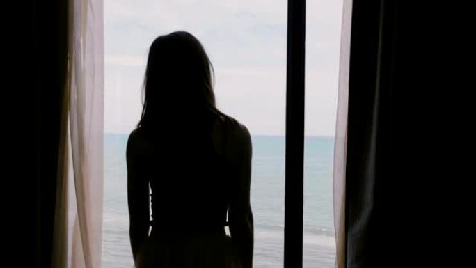 年轻快乐的女人在黑暗的房间里打开窗帘，欣赏令人难以置信的海浪景色，触摸头发慢动作。
