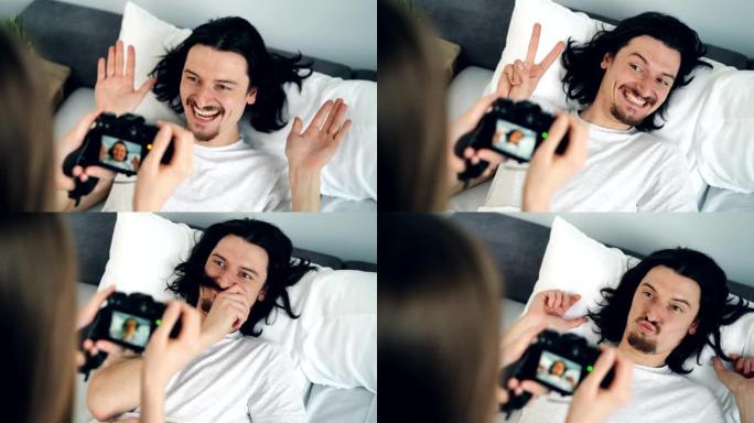 富有创造力的男人在床上摆出滑稽的面孔，而女人用相机拍照
