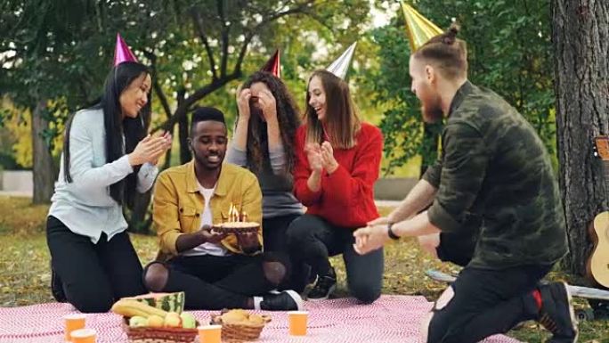 有爱心的朋友将蛋糕带给坐在公园野餐的毯子上的非洲裔美国人，闭着眼睛，他吹着蜡烛，笑着享受惊喜。
