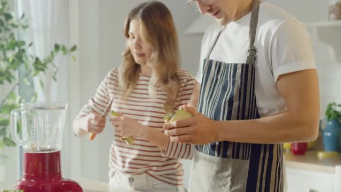 戴着围裙的英俊年轻男子和漂亮的女孩正在厨房里做冰沙。幸福的夫妇正在准备健康的有机饮料。晴天在家男女。