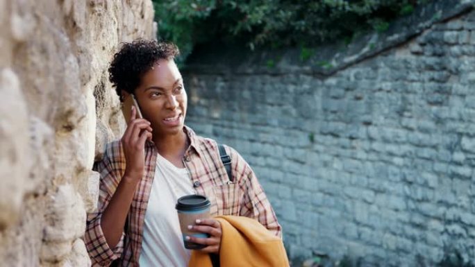 千禧一代黑人女性，短卷发，穿着格子衬衫，靠在石墙上，用智能手机聊天，喝外卖咖啡，选择性聚焦