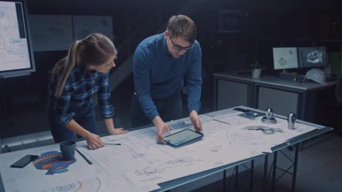 在黑暗的工业设计工程设施中，男女工程师使用数字平板电脑和会议桌在蓝图上交谈和工作。在桌面图纸和发动机