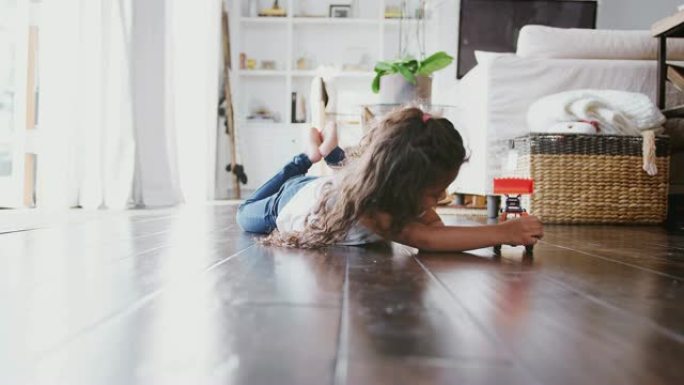 年轻的西班牙裔女孩躺在客厅的地板上玩玩具挖掘机卡车，前视图