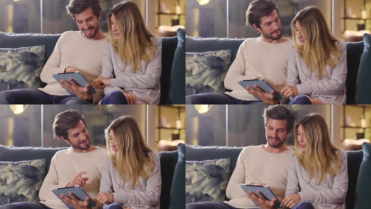 一对夫妇在沙发上的客厅里看着平板电脑，上面放着他们假期的纪念品照片，并开心地微笑着。