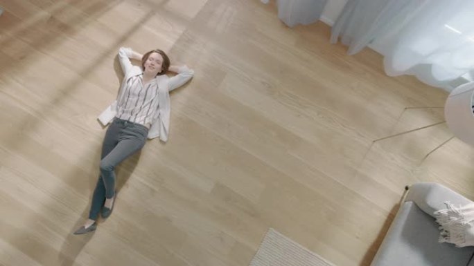 年轻女子躺在公寓的木地板上。她穿得很随意。舒适的客厅配有现代简约的内部和木质镶木地板。顶视图摄像机镜