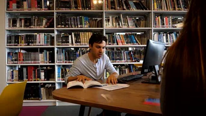 年轻的男学生在图书馆的计算机上工作时看着一些文件和一本书