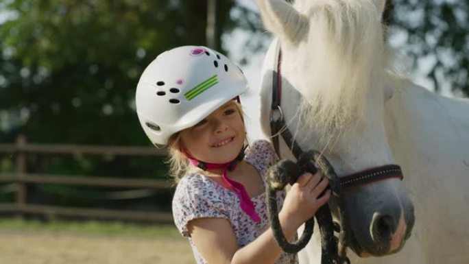 一个戴着骑师头盔的可爱的小女孩的真实镜头正在阳光下的马stable上爱抚着一匹白色的小马