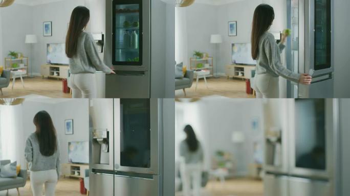 穿着白色牛仔裤和灰色毛衣的美丽黑发女孩来到带玻璃门的高科技冰箱，拿着一个青苹果。她去客厅。公寓有现代