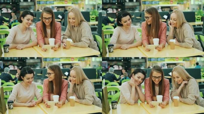 快乐的年轻女士们一起使用智能手机，看着屏幕微笑和大笑，坐在购物中心的咖啡馆里，可见纸袋。小玩意和有趣