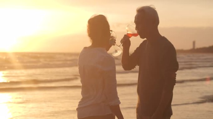 浪漫的亚洲老年夫妇在美丽的热带海滩上慢动作观看日落，享受一杯葡萄酒。