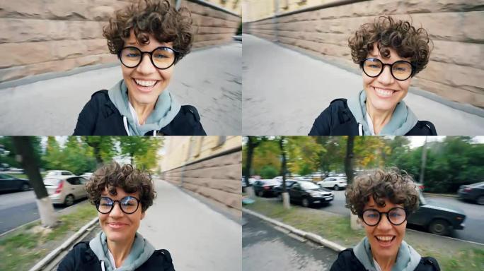 无忧无虑的年轻女士的视角镜头，短发和眼镜看着相机，在秋天的日子里在街上开心地笑着转身。