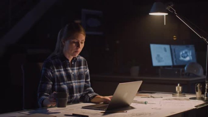 坐在办公桌前的漂亮女工程师在笔记本电脑上工作。躺在桌子上的蓝图。在黑暗的工业设计工程设施中。暖灯