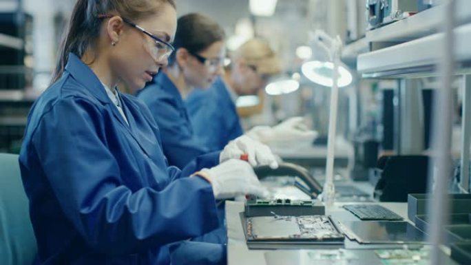 穿着蓝色工作服和防护眼镜的电子工厂女工正在用螺丝刀组装笔记本电脑的主板。拥有多名员工的高科技工厂设施