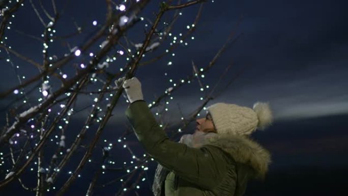 女士在户外树上悬挂圣诞装饰品，并带有串灯