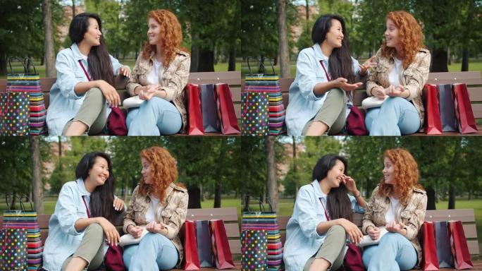 美丽的亚洲和高加索女孩在公园里用购物袋坐在长凳上大笑