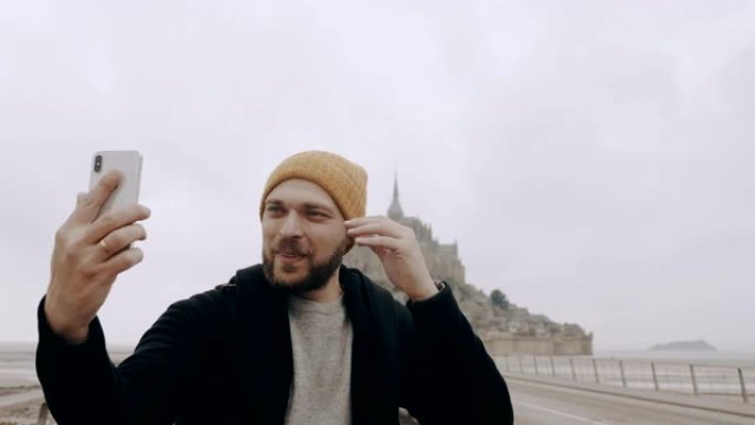 快乐的年轻兴奋的高加索博客男子拍摄史诗圣米歇尔山城堡慢动作的智能手机自拍视频。