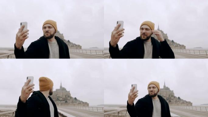 快乐的年轻兴奋的高加索博客男子拍摄史诗圣米歇尔山城堡慢动作的智能手机自拍视频。