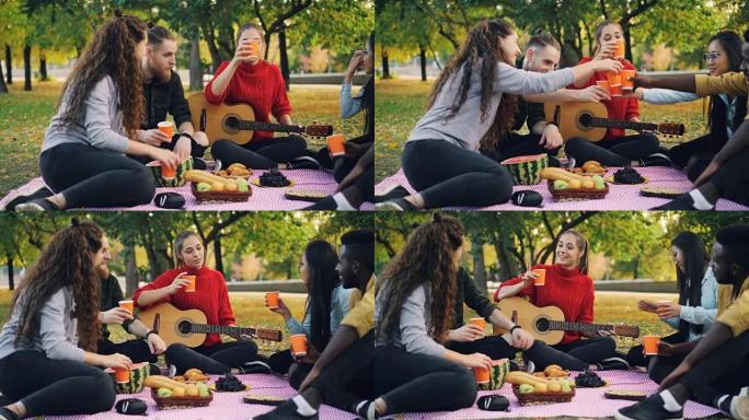 在温暖的秋天，年轻的男人和女人在公园野餐时用吉他敬酒和碰杯。友谊、饮料和休闲活动理念。