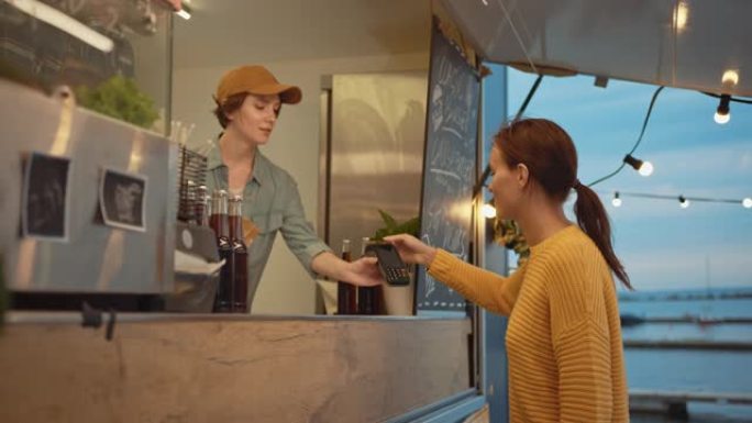 Food Truck员工将新鲜制作的汉堡交给一个快乐的年轻女性。年轻女士正在用非接触式信用卡支付食物
