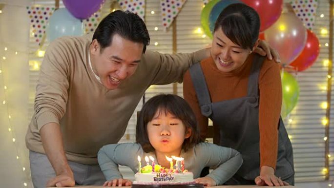 一个可爱的亚洲小女孩坐在餐桌旁，在生日蛋糕上吹蜡烛，而她的家人站在后面，为她唱歌。她露出灿烂的笑容，