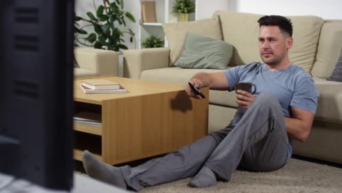 男人坐在地板上喝茶，一边在家看电视