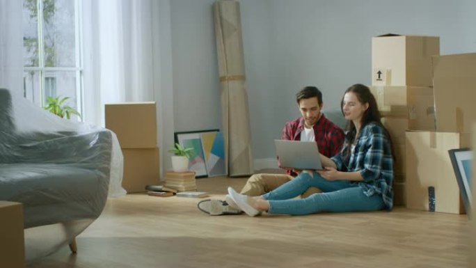 幸福的年轻夫妇坐在新租/购买的公寓的地板上使用笔记本电脑。在现代明亮的甜蜜家庭中打开包装的纸板箱和有