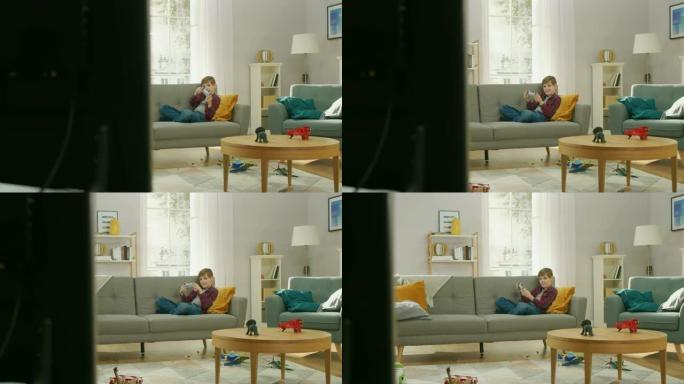 可爱的小男孩躺在沙发上，使用操纵杆控制器在电视控制台上玩视频游戏。男孩在家里玩电子游戏。