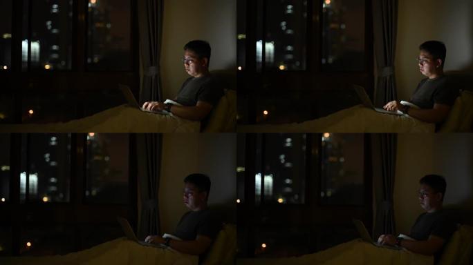 一个亚洲华人中年人晚上在家工作，在床上用他的笔记本电脑
