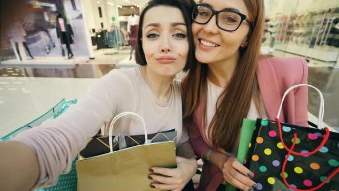年轻女学生在购物中心自拍的视点镜头，展示明亮的纸袋和竖起大拇指，做鬼脸。购物狂和快乐概念。