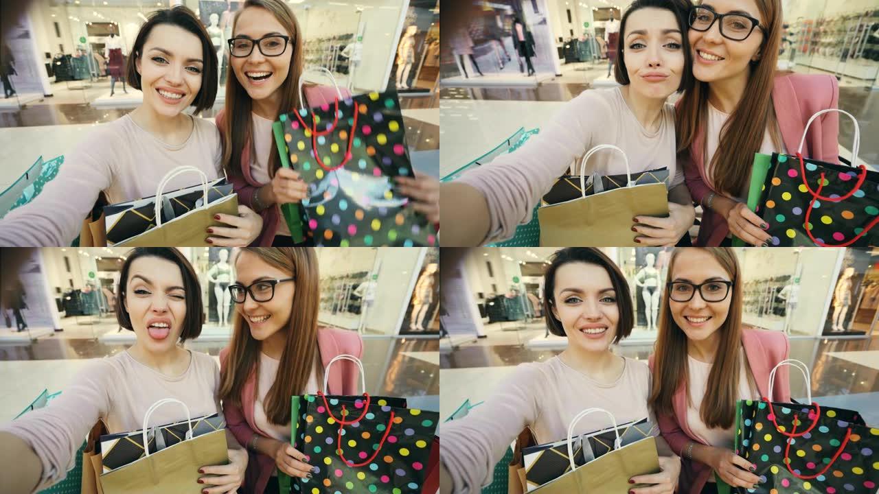 年轻女学生在购物中心自拍的视点镜头，展示明亮的纸袋和竖起大拇指，做鬼脸。购物狂和快乐概念。