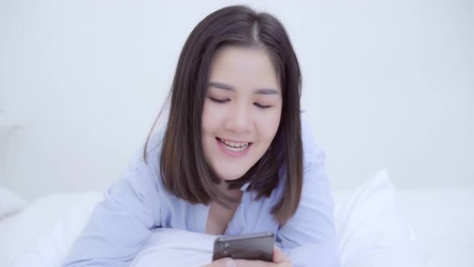 年轻的亚洲女性在早上醒来后躺在床上使用智能手机，美丽迷人的日本女孩在家里的卧室里微笑放松。享受时间生