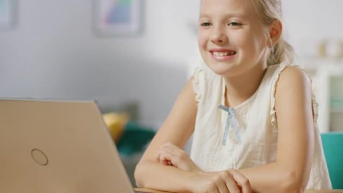 可爱的小女孩坐在客厅的桌子旁使用笔记本电脑。孩子在浏览互联网并观看卡通和有趣的视频时很有趣。