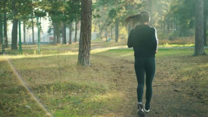 穿着运动服的苗条年轻女子在公园慢跑，戴着耳机独自听音乐训练。轻柔、运动和自然概念。