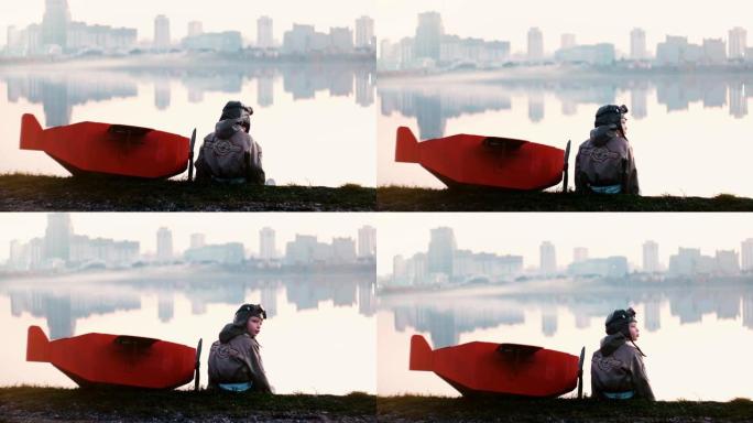 穿着旧飞行员服装的小男孩坐在神奇的城市湖附近，穿着有趣的红色纸板飞机慢动作。