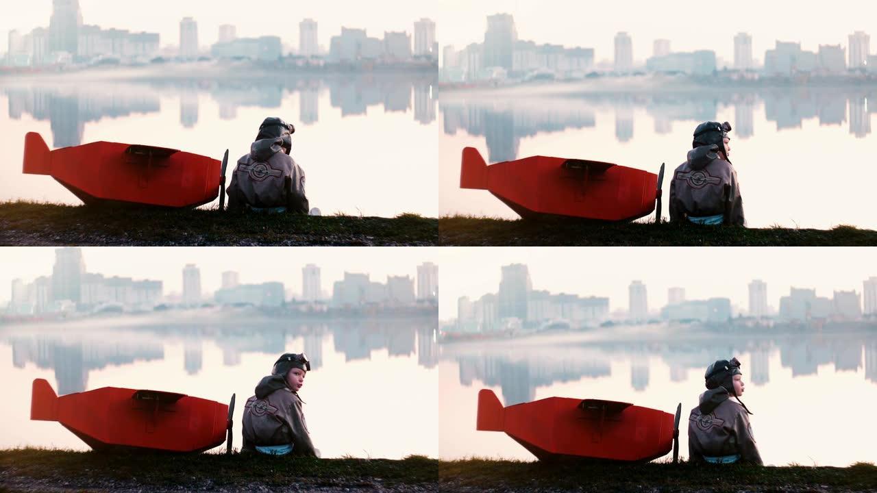穿着旧飞行员服装的小男孩坐在神奇的城市湖附近，穿着有趣的红色纸板飞机慢动作。
